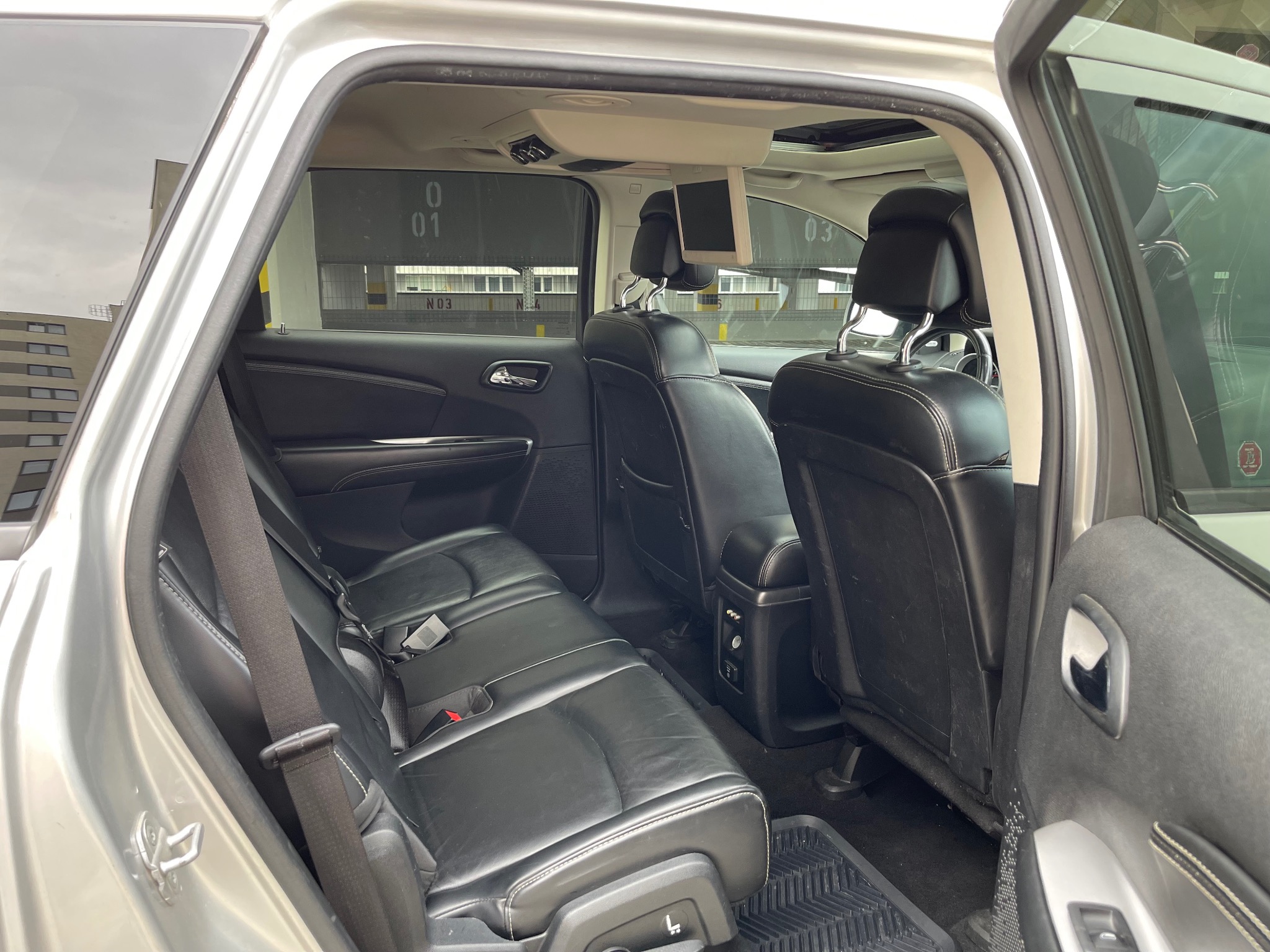 Wnętrze samochodu z czarną skórzaną tapicerką, widok na tylne siedzenia z zamontowanymi ekranami w zagłówkach i otwartymi drzwiami ukazującymi budynek w tle.