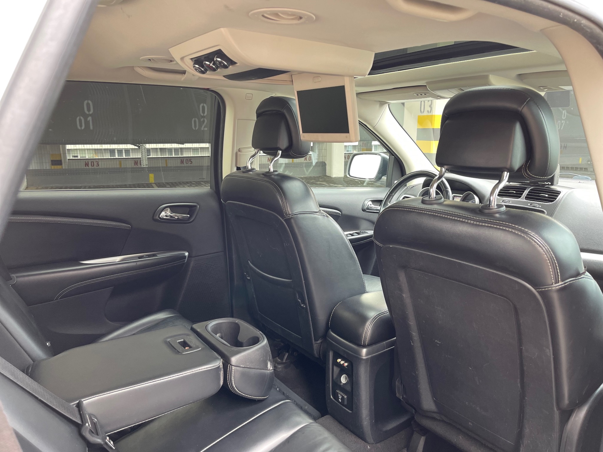 Wnętrze czarnego samochodu z skórzanymi siedzeniami, widok z tyłu z rozkładanym podłokietnikiem i uchwytami na napoje, ekran zamontowany na suficie, widok na parking przez przyciemnione okno.