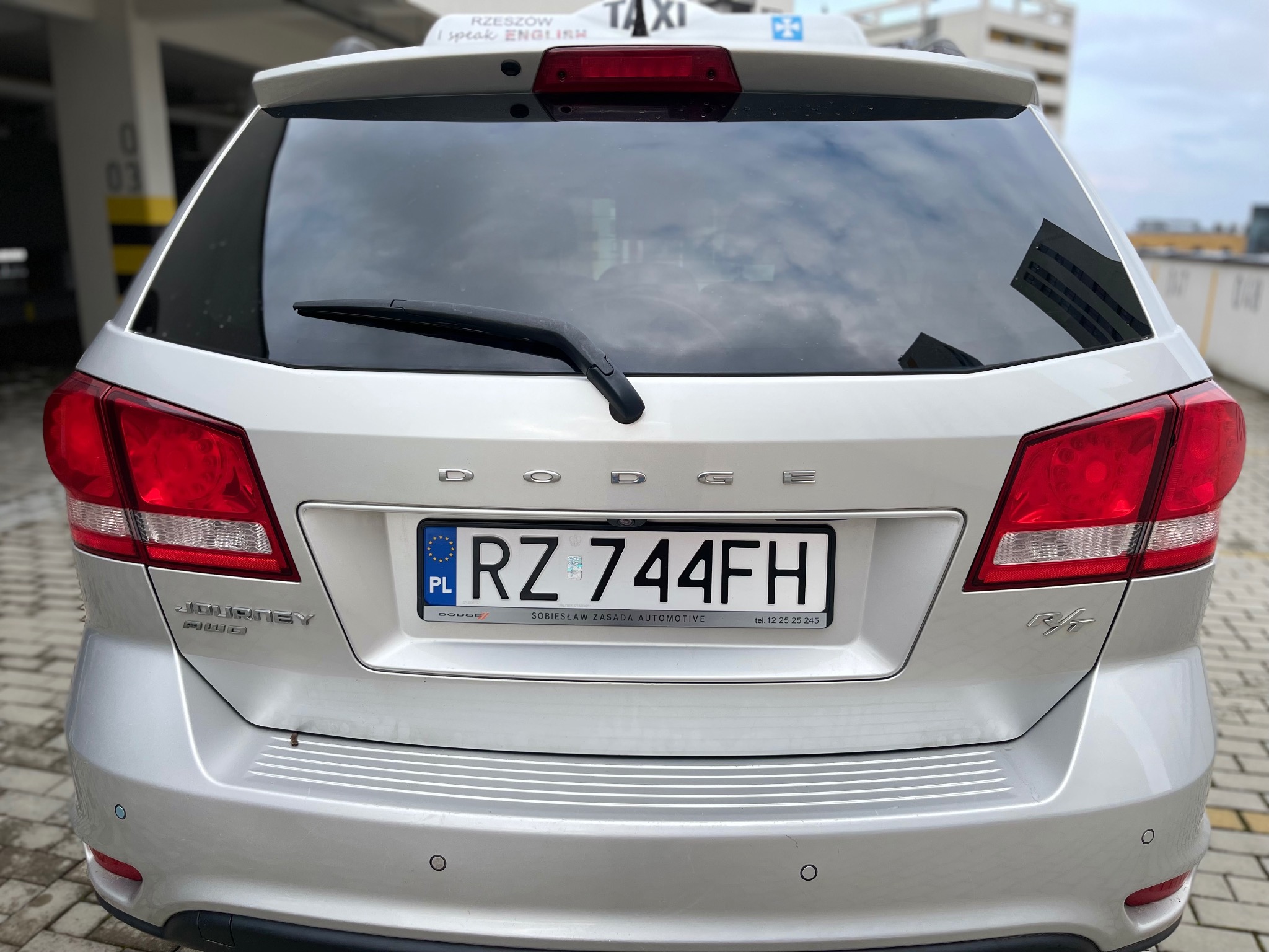 Tył srebrnego samochodu Dodge Journey z tablicą rejestracyjną RZ 744FH, oznaczony jako taksówka z napisem 'Rzeszów I speak ENGLISH' na dachu, zaparkowany w garażu podziemnym.