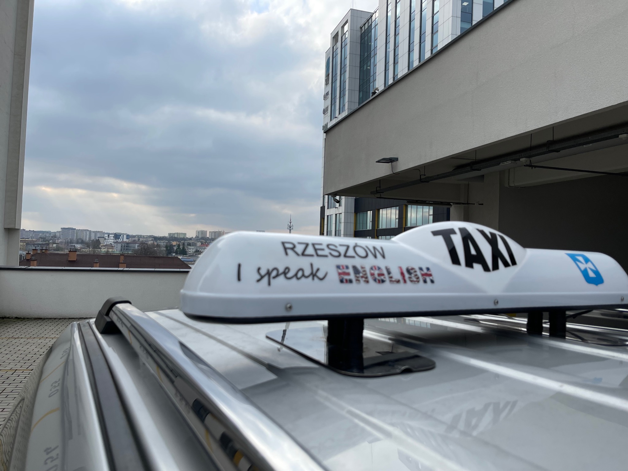 Lampa taksówkowa na dachu srebrnego samochodu z napisem 'Rzeszów I speak ENGLISH' i logo taksówki na tle miejskiego krajobrazu i nowoczesnego budynku.