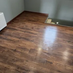 Ułożone panele podłogowe w odcieniach brązu, widoczny fragment grzejnika i okno z parapetem, brak listew przypodłogowych.