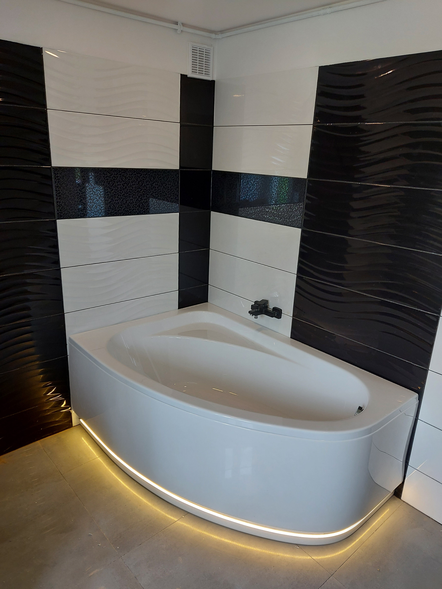 Nowoczesna łazienka z wanną narożną podświetloną taśmą LED, ściany wyłożone czarno-białymi płytkami z falistym wzorem.