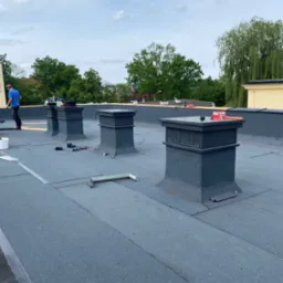 Prace na dachu pokrytym papą, widoczne cztery kominy z szarymi obudowami, narzędzia, materiały budowlane i pracownik w niebieskiej koszulce w tle.