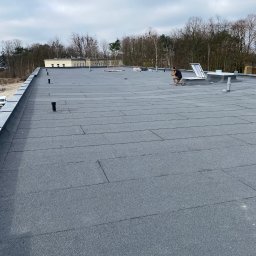 MK Marek Kuznowicz - Dekarz pracujący na płaskim dachu pokrytym papą termozgrzewalną, w tle budynek i drzewa.