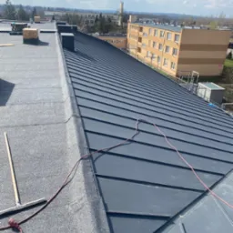 Widok z góry na fragment dachu pokrytego papą i blachą trapezową w kolorze grafitowym, z widocznym kablem zasilającym i narzędziem do rozprowadzania masy, budynki mieszkalne w tle.