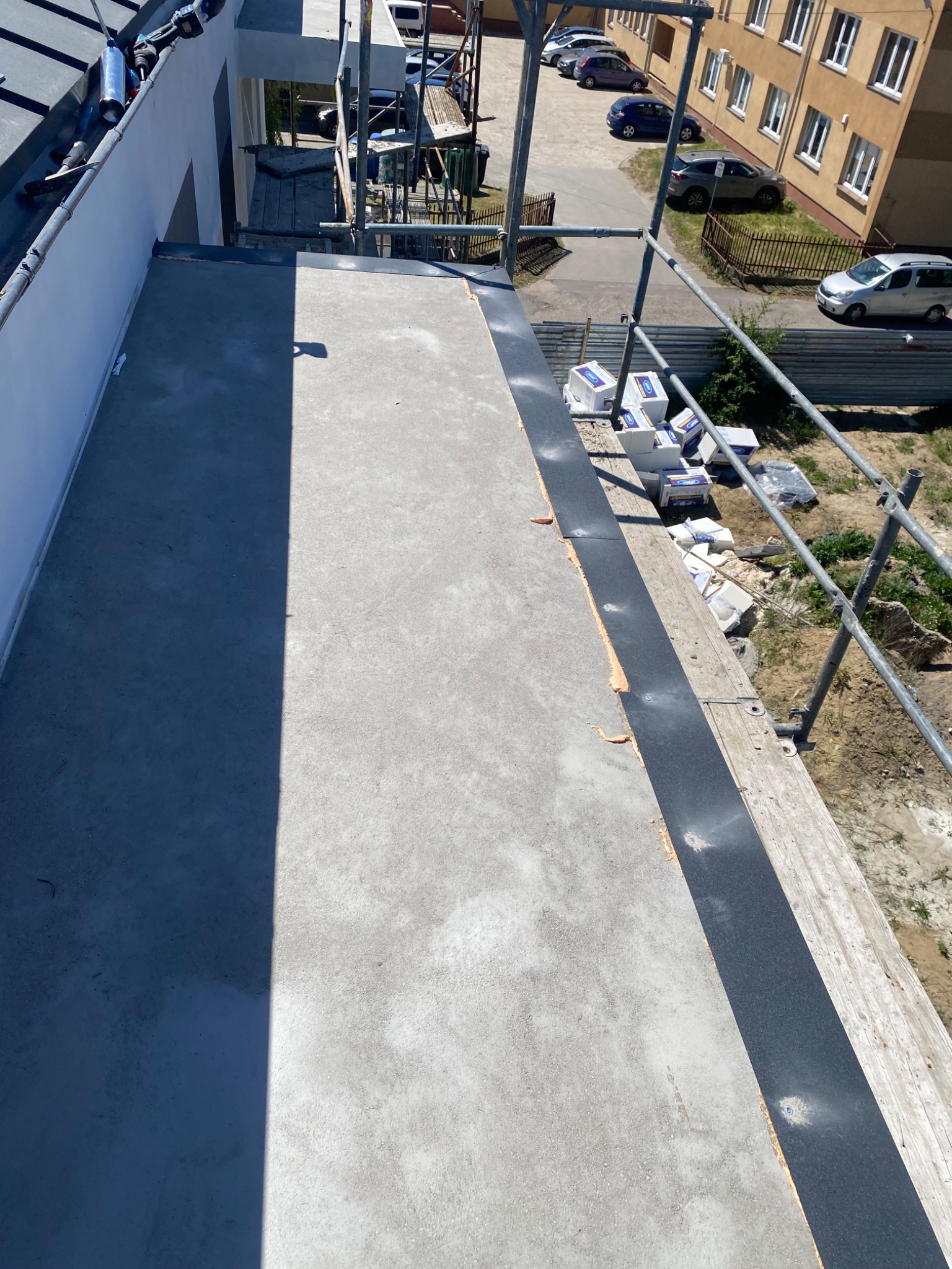 Widok z góry na płaski dach w trakcie prac izolacyjnych, z widocznymi materiałami izolacyjnymi i rusztowaniem w tle.