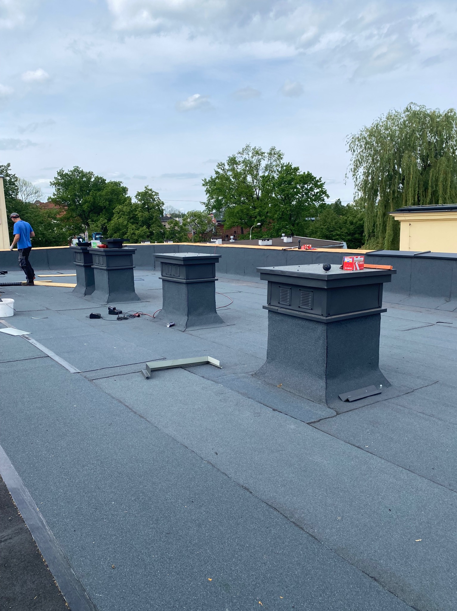 Prace na dachu pokrytym papą, widoczne cztery kominy z szarymi obudowami, narzędzia, materiały budowlane i pracownik w niebieskiej koszulce w tle.