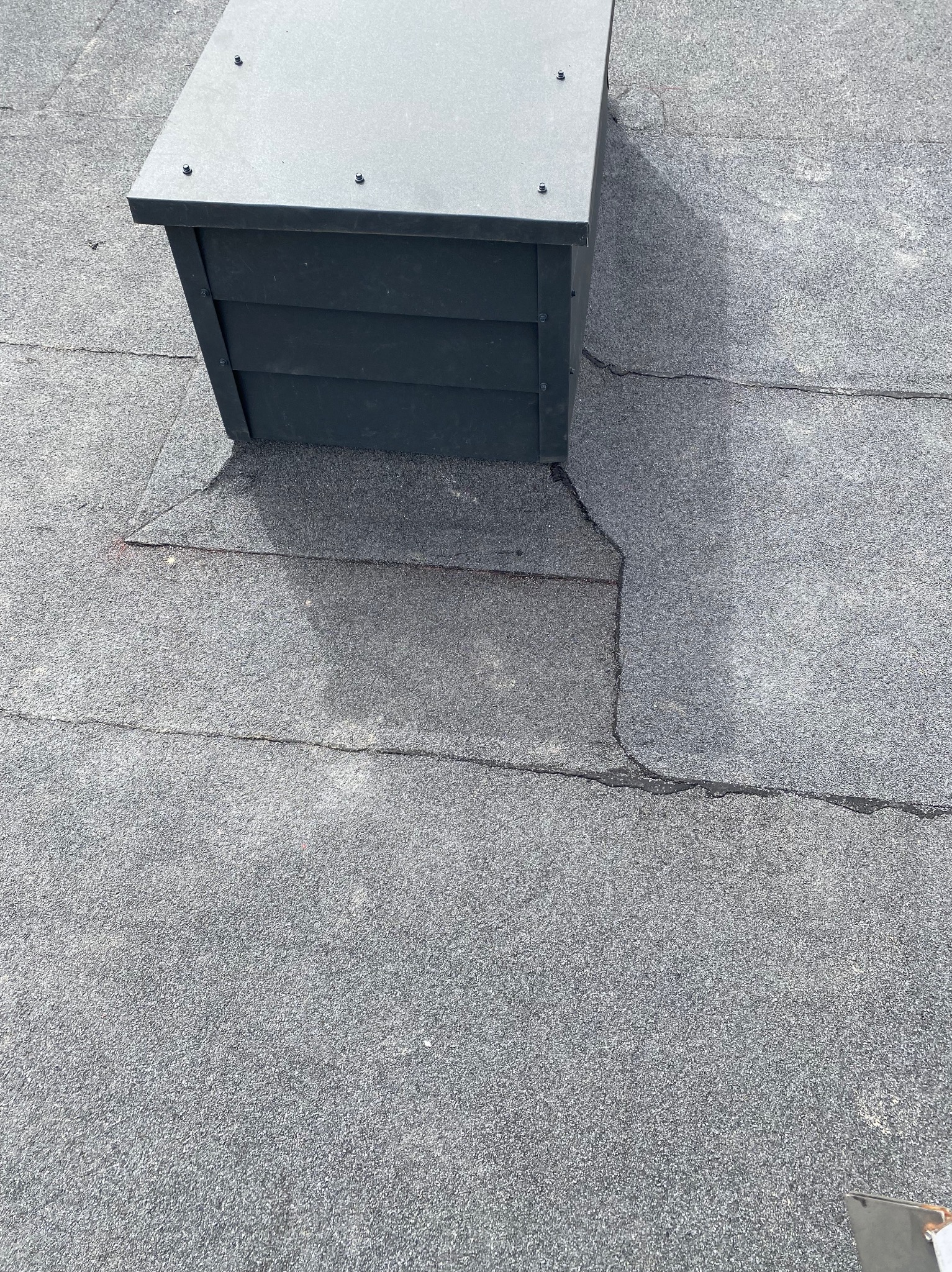 Szczegółowe ujęcie fragmentu dachu pokrytego papą z kominkiem wentylacyjnym w kształcie domku z szarej blachy, widoczne łączenia papy i cień kominka.