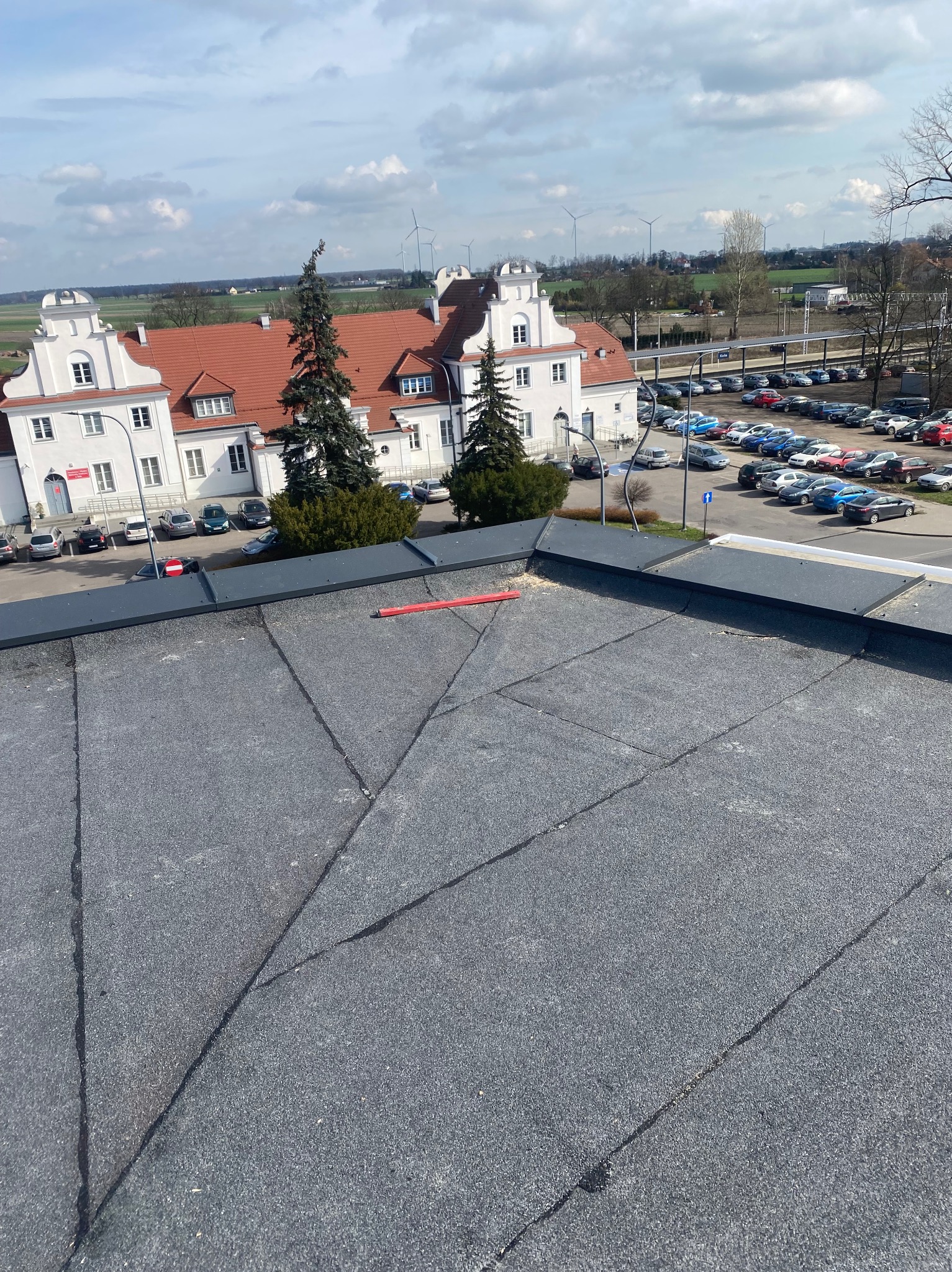 Widok z góry na płaski dach pokryty papą, z budynkiem w tle i parkingiem pełnym samochodów