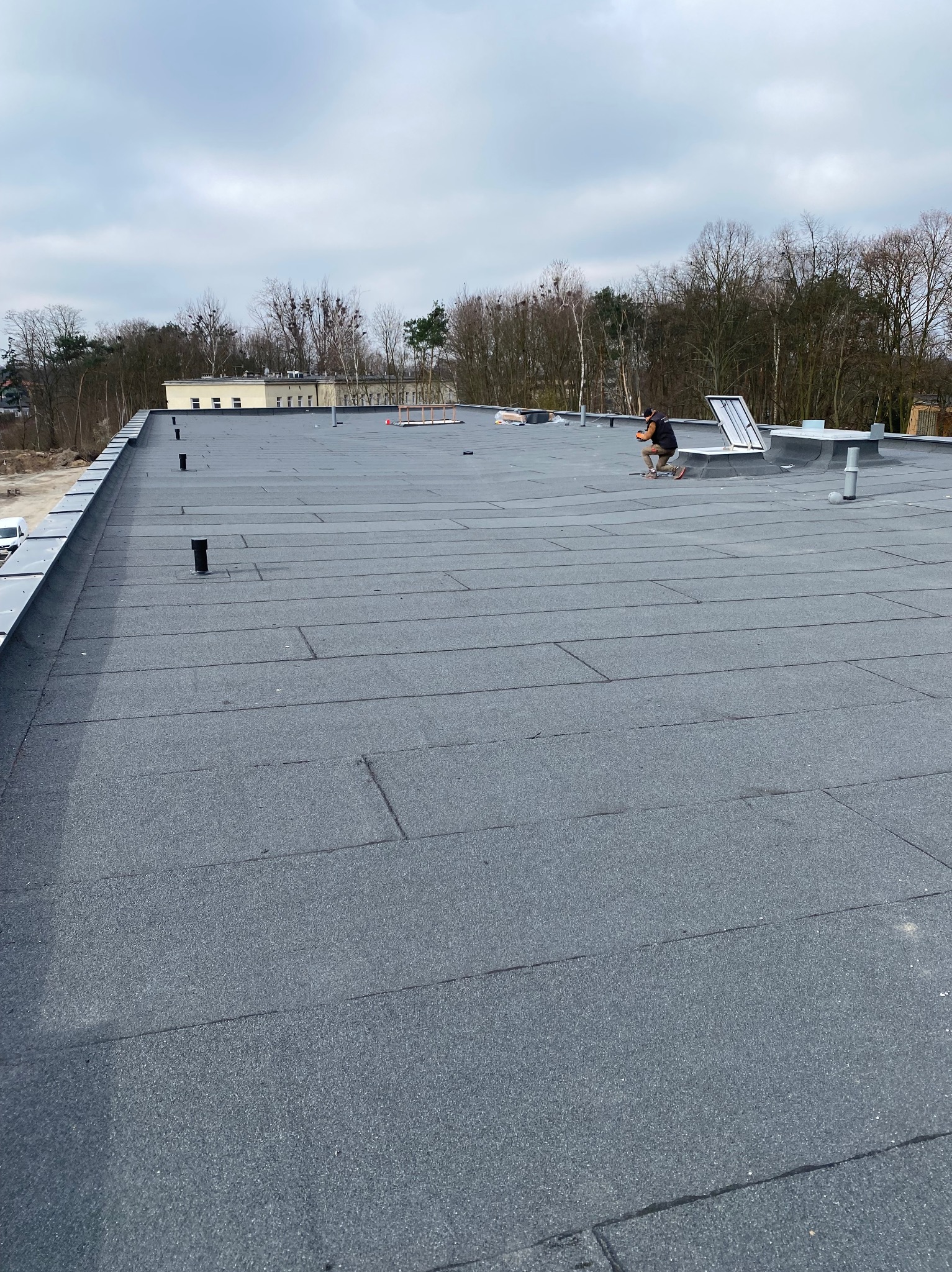 Dekarz pracujący na płaskim dachu pokrytym papą termozgrzewalną, w tle budynek i drzewa.