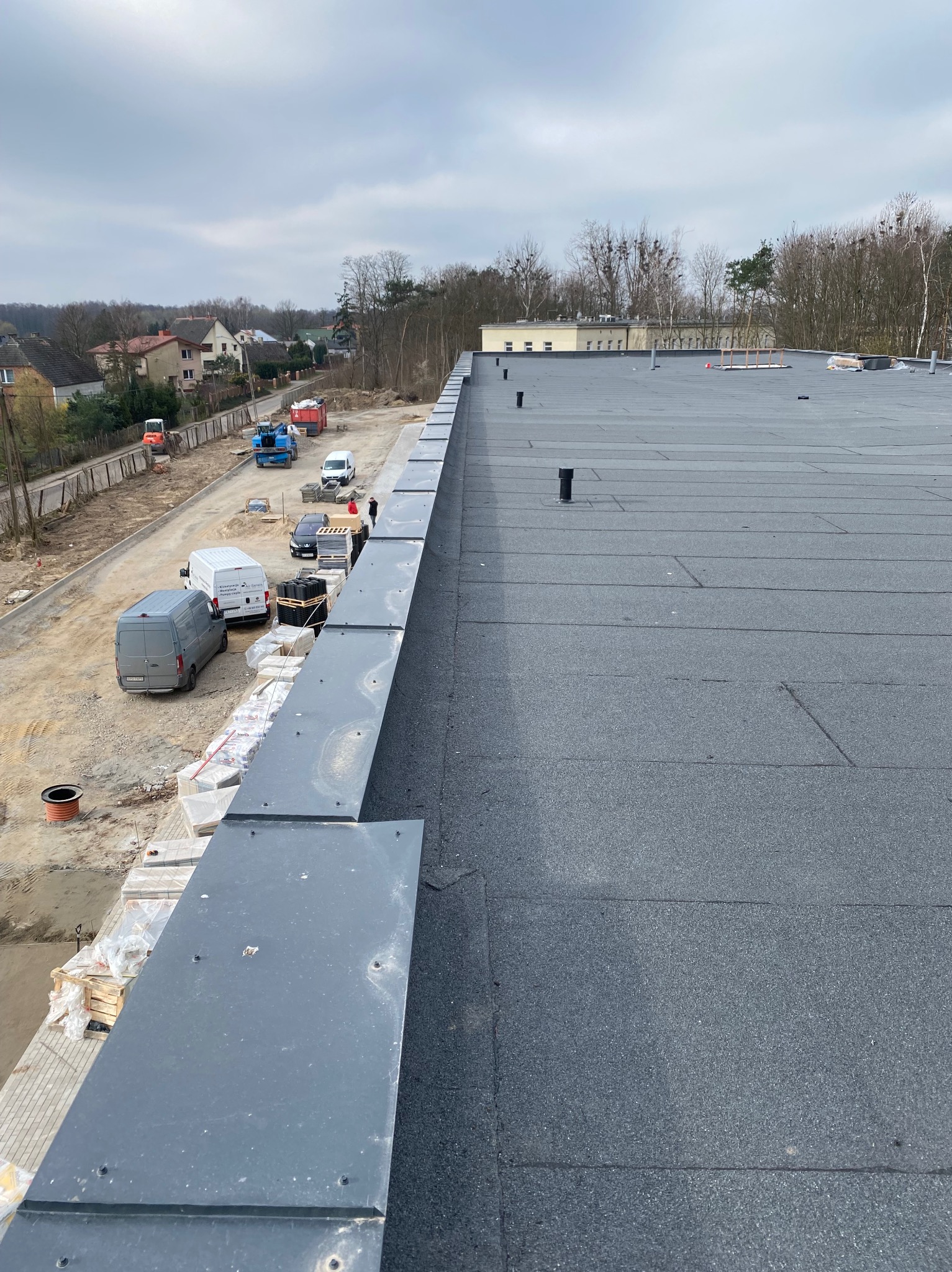 Widok z góry na nowo pokryty, płaski dach z papy termozgrzewalnej, z widocznymi kominkami wentylacyjnymi i obróbkami blacharskimi na krawędzi, w tle zaparkowane samochody i budynki mieszkalne.