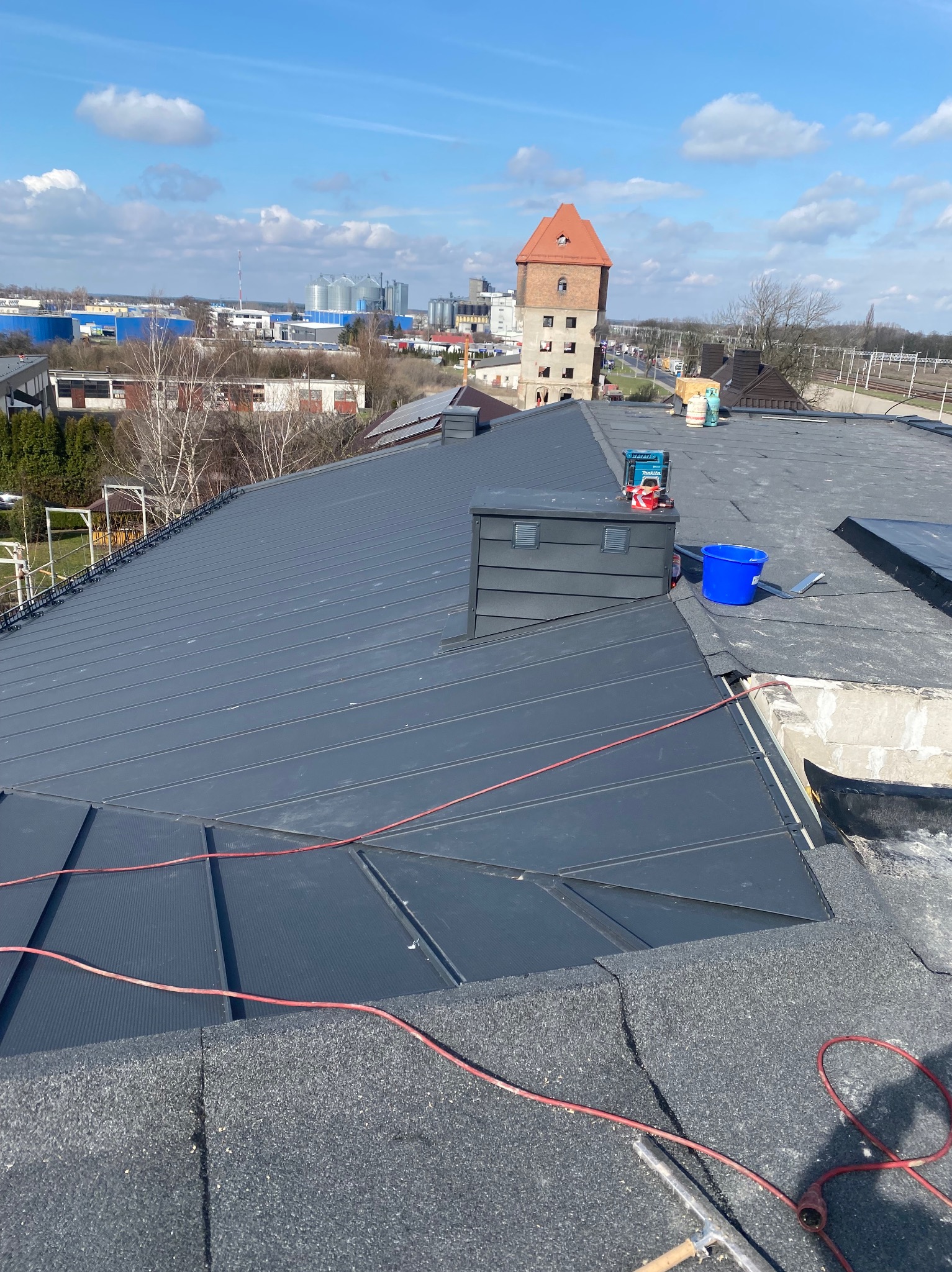 Nowoczesny, grafitowy dach z blachy na tle miejskiego krajobrazu z wieżą i silosami w oddali, widoczne narzędzia i akcesoria dekarskie podczas prac.