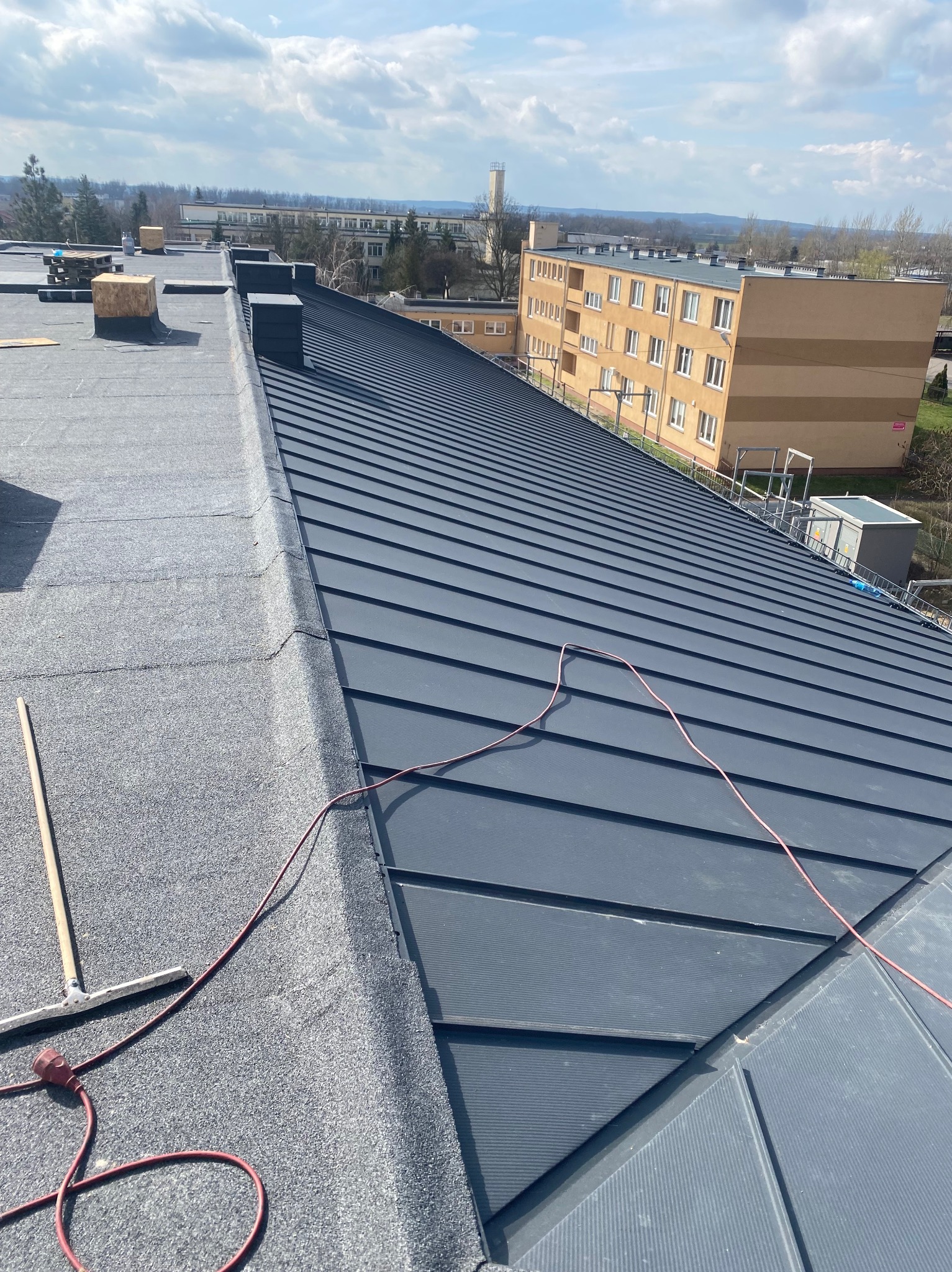 Widok z góry na fragment dachu pokrytego papą i blachą trapezową w kolorze grafitowym, z widocznym kablem zasilającym i narzędziem do rozprowadzania masy, budynki mieszkalne w tle.