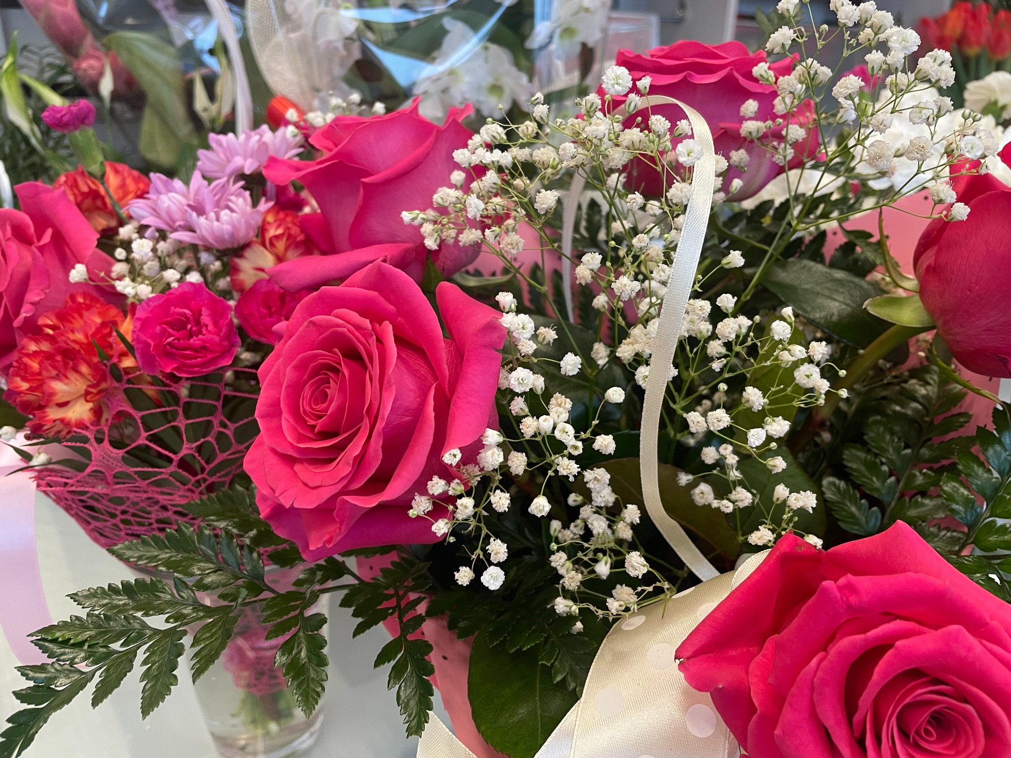 Bukiet mieszanych kwiatów w odcieniach różu, czerwieni i bieli, z dominującymi różami i drobnymi białymi kwiatami, przewiązany wstążką w kropki.