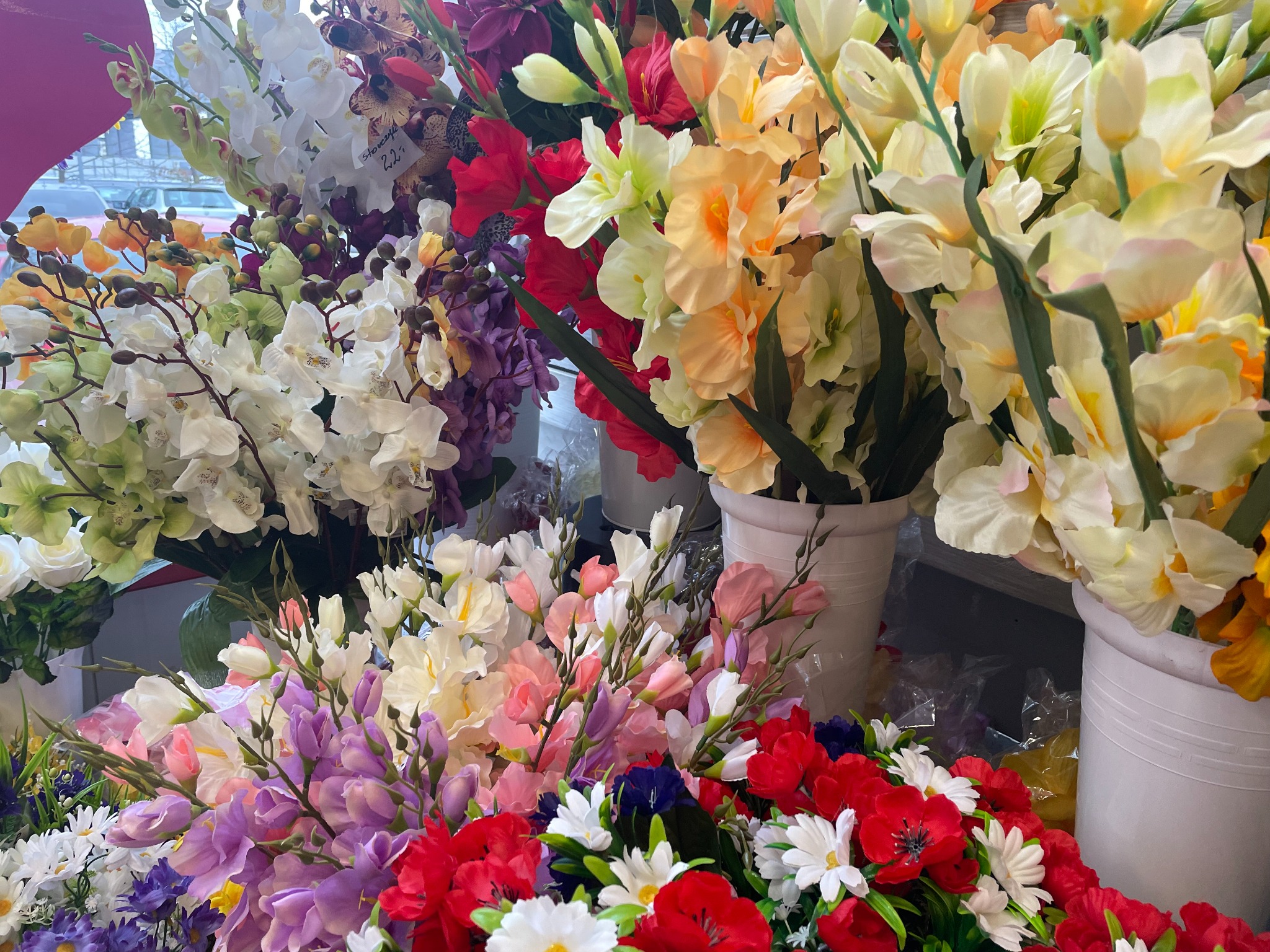 Różnorodny asortyment sztucznych kwiatów w odcieniach bieli, czerwieni, fioletu i żółci, ustawionych w białych donicach, widoczna cena na jednym z bukietów.