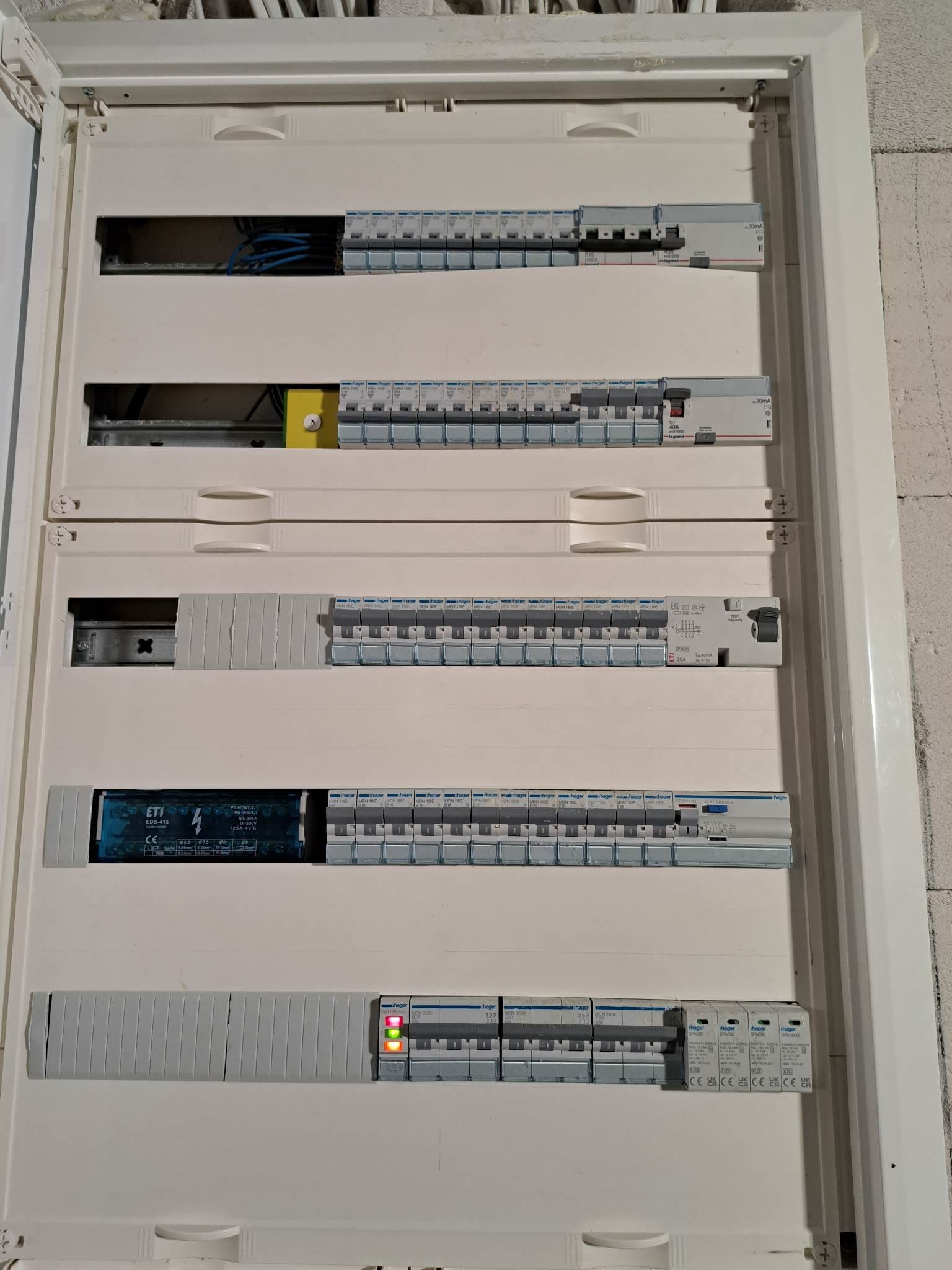 Otwarta, biała skrzynka rozdzielcza z widocznymi bezpiecznikami, wyłącznikami różnicowoprądowymi marek Hager i Legrand oraz przewodami elektrycznymi, z trzema zapalonymi diodami sygnalizacyjnymi...