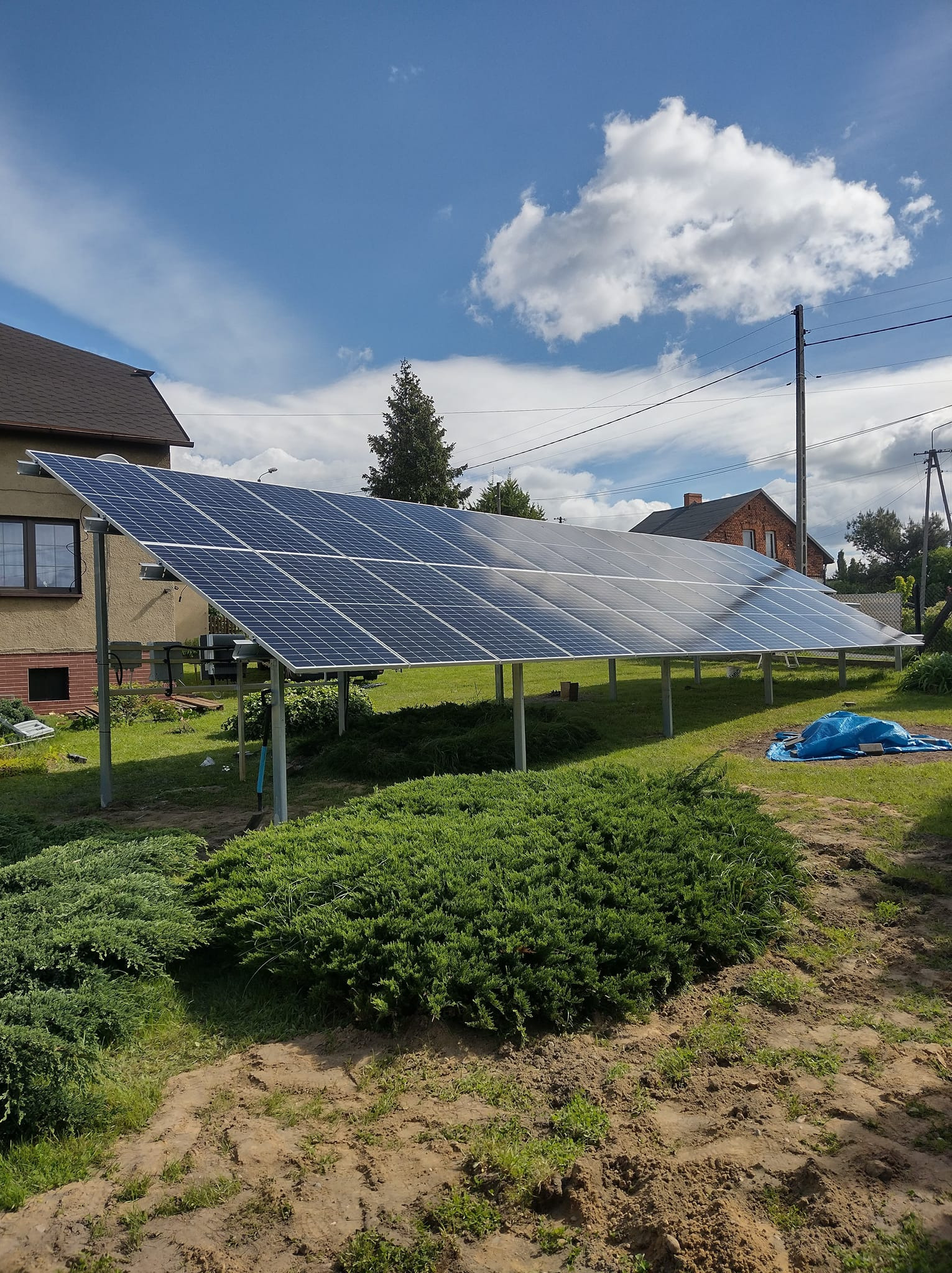 Instalacja paneli słonecznych na metalowej konstrukcji w ogrodzie, widoczny dom z brązowym dachem, niebieska plandeka i częściowo zachmurzone niebo.