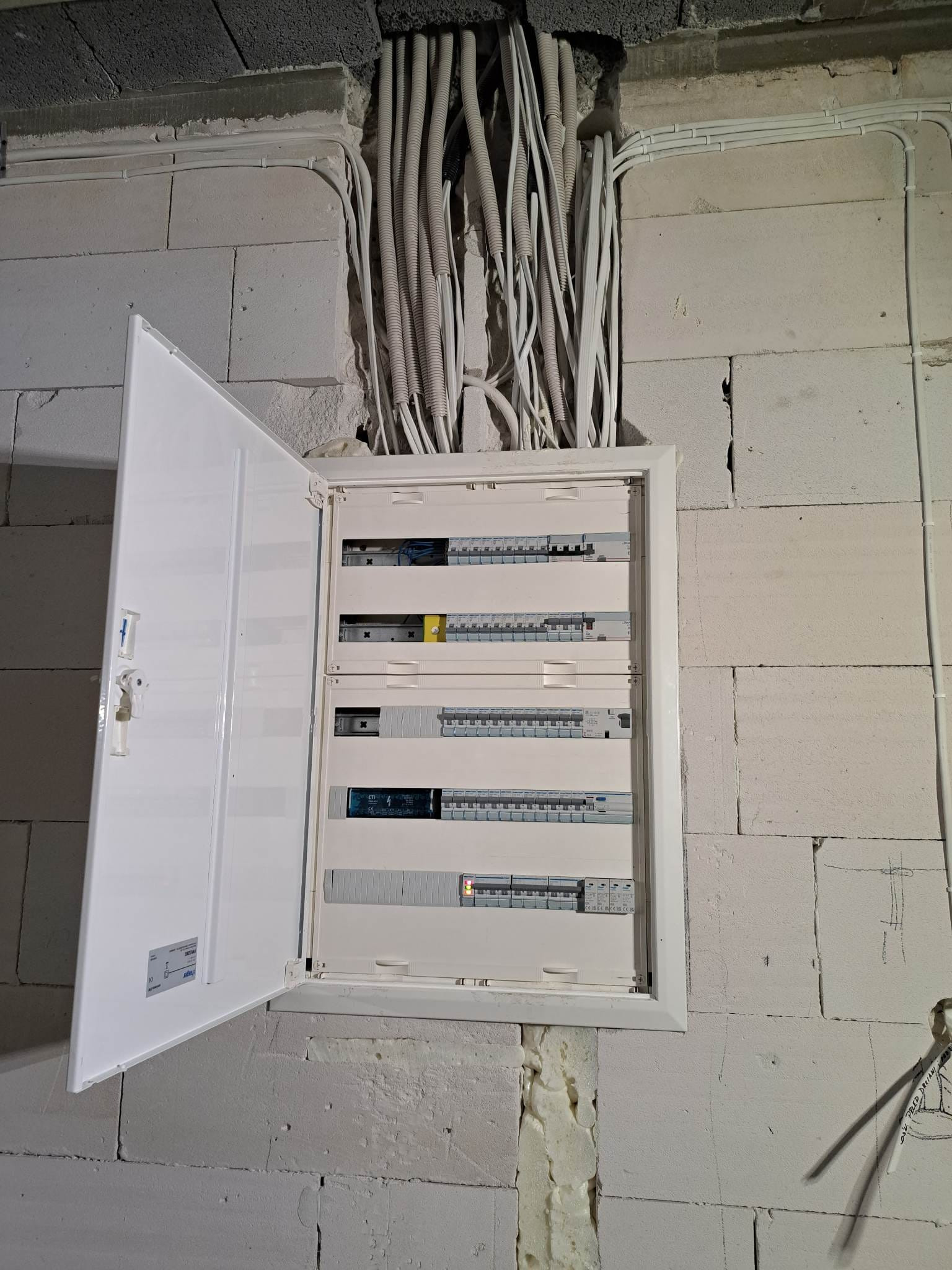 Otwarta skrzynka elektryczna z bezpiecznikami zamontowana na ścianie z betonu komórkowego, widoczne kable w peszlach wchodzące do skrzynki od góry.