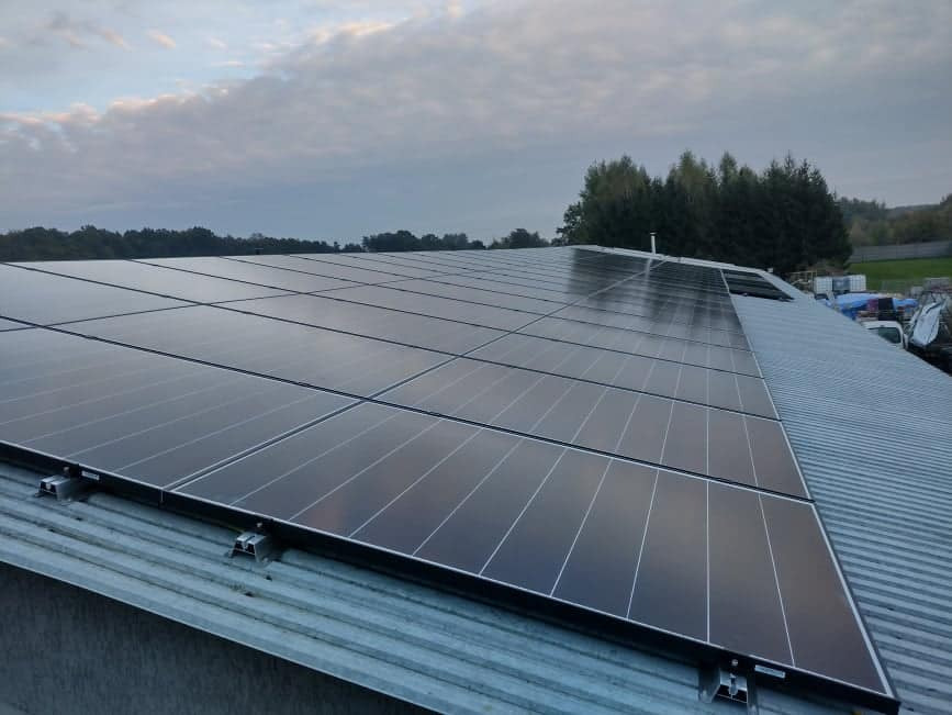 Panel słoneczny na metalowym dachu, widoczne mocowania, pochmurne niebo w tle.