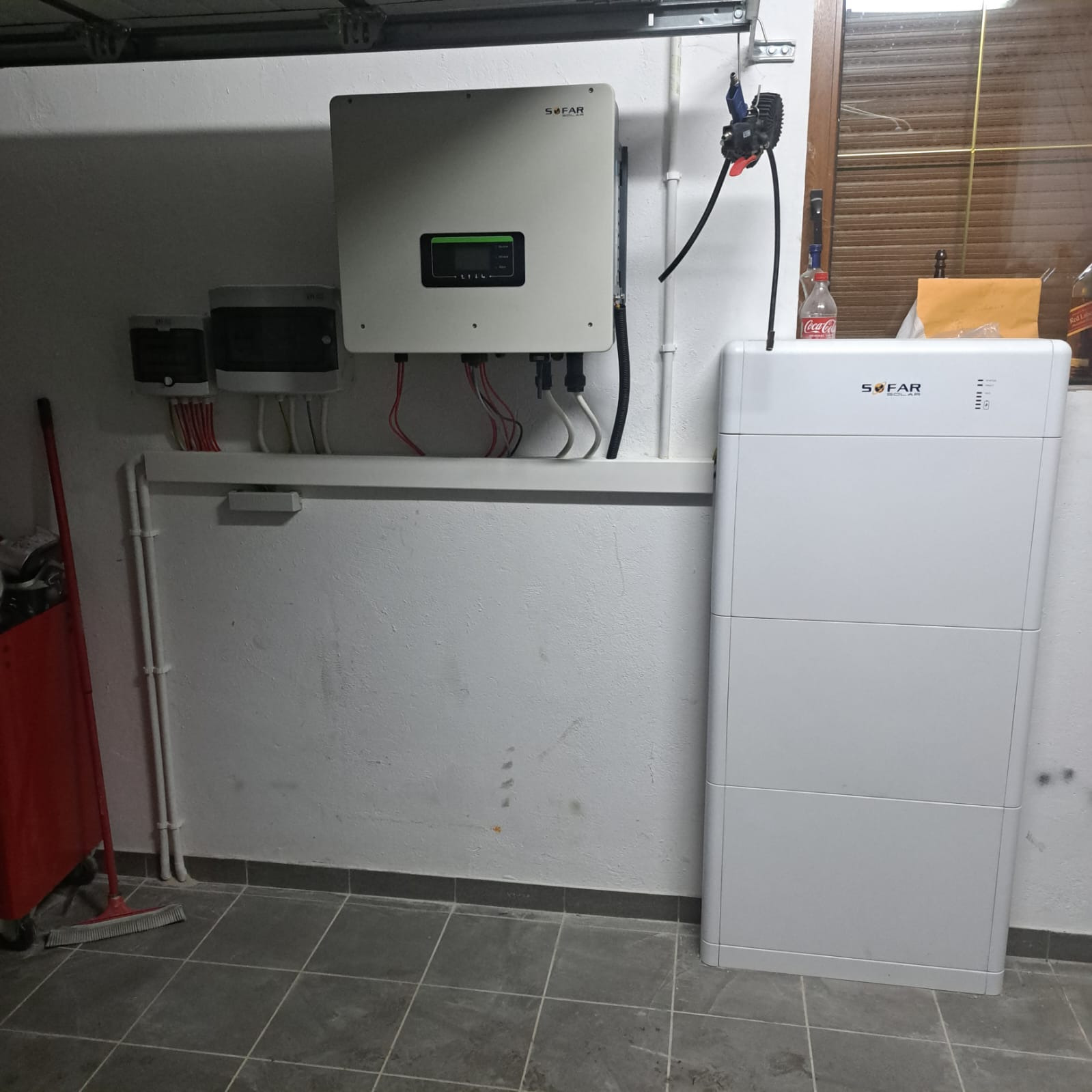 Biała bateria solarna SOFAR, falownik i skrzynki elektryczne zamontowane na pomalowanej ścianie w garażu, widoczne przewody i rurki instalacyjne.