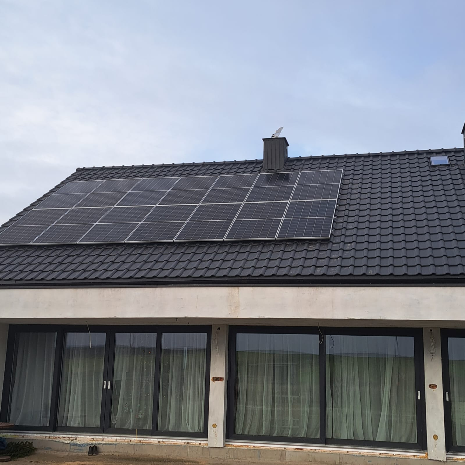 Panele fotowoltaiczne zamontowane na dachu domu z czarną dachówką, widoczne kominy i okno dachowe. Dom w stanie surowym otwartym z dużymi oknami.