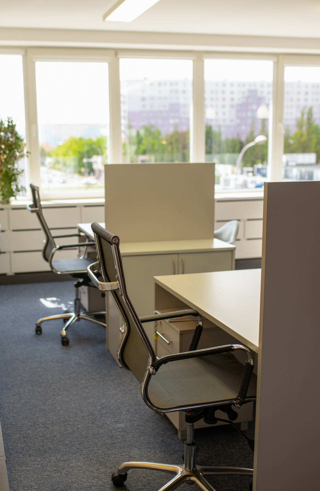 Jasne biuro z dwoma stanowiskami pracy: biurka z przegrodami, ergonomiczne krzesła obrotowe z siatkowym oparciem, widok na miasto za oknem.
