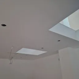 Biały sufit z dwoma świetlikami, widoczne otwory na oświetlenie i wystające kable elektryczne w nowo wykończonym pomieszczeniu.