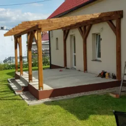Drewniana pergola zadaszająca betonowy taras przy domu z czerwoną dachówką, widoczne narzędzia i drabina, gniazdo bocianie na słupie.