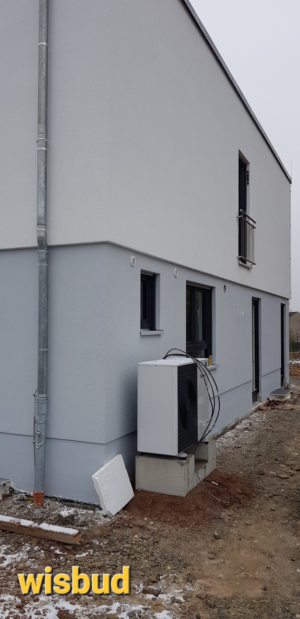Dwukondygnacyjny budynek z szarą elewacją, widoczny fragment ogrodu z pompą ciepła na betonowym fundamencie, rura spustowa i fragment styropianu na ziemi.