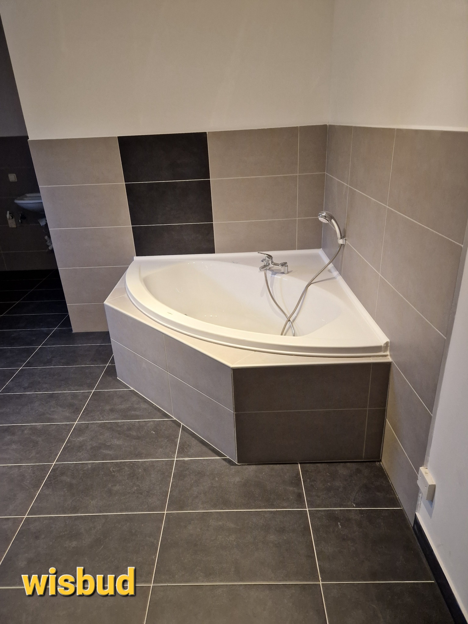 Narożna wanna obudowana szarymi płytkami w łazience z ciemną podłogą i jasnymi ścianami, widoczna bateria i słuchawka prysznicowa.