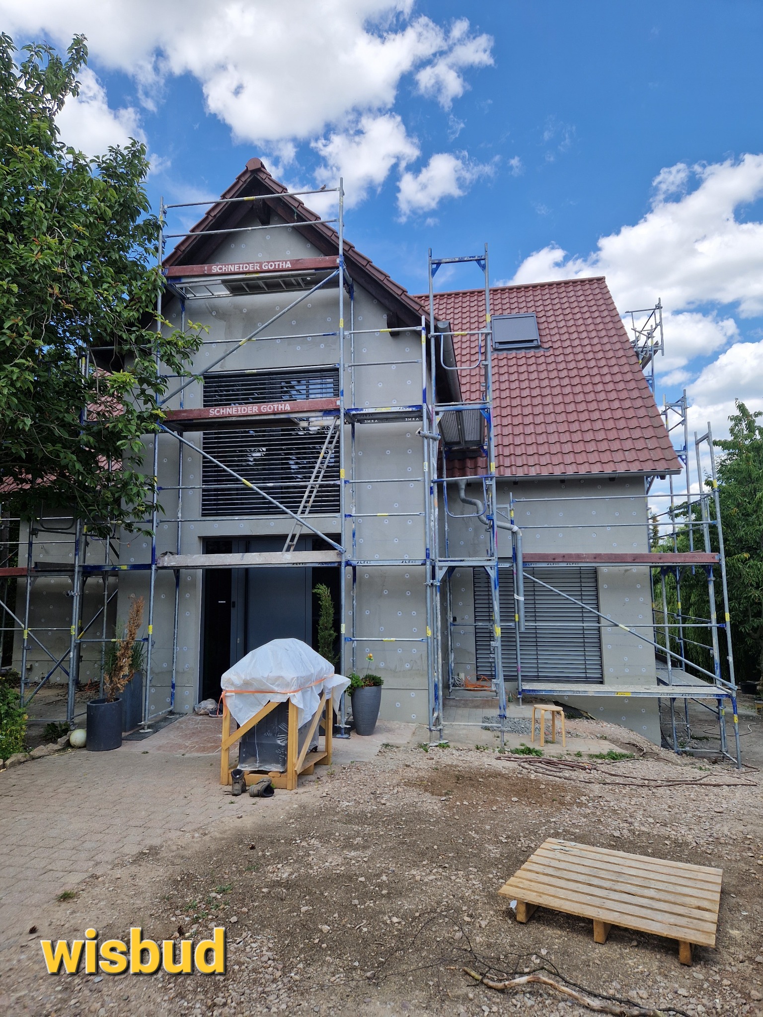 Dom w trakcie ocieplania elewacji, widoczne rusztowania, styropianowe płyty z zaślepkami, częściowo zakryte okna i dach z czerwonej dachówki.