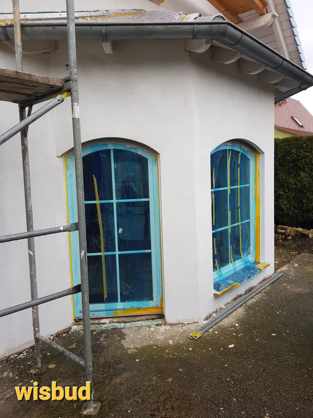 Biała elewacja budynku w trakcie prac, okna zabezpieczone folią i taśmą, widoczne rusztowanie i fragment dachu.