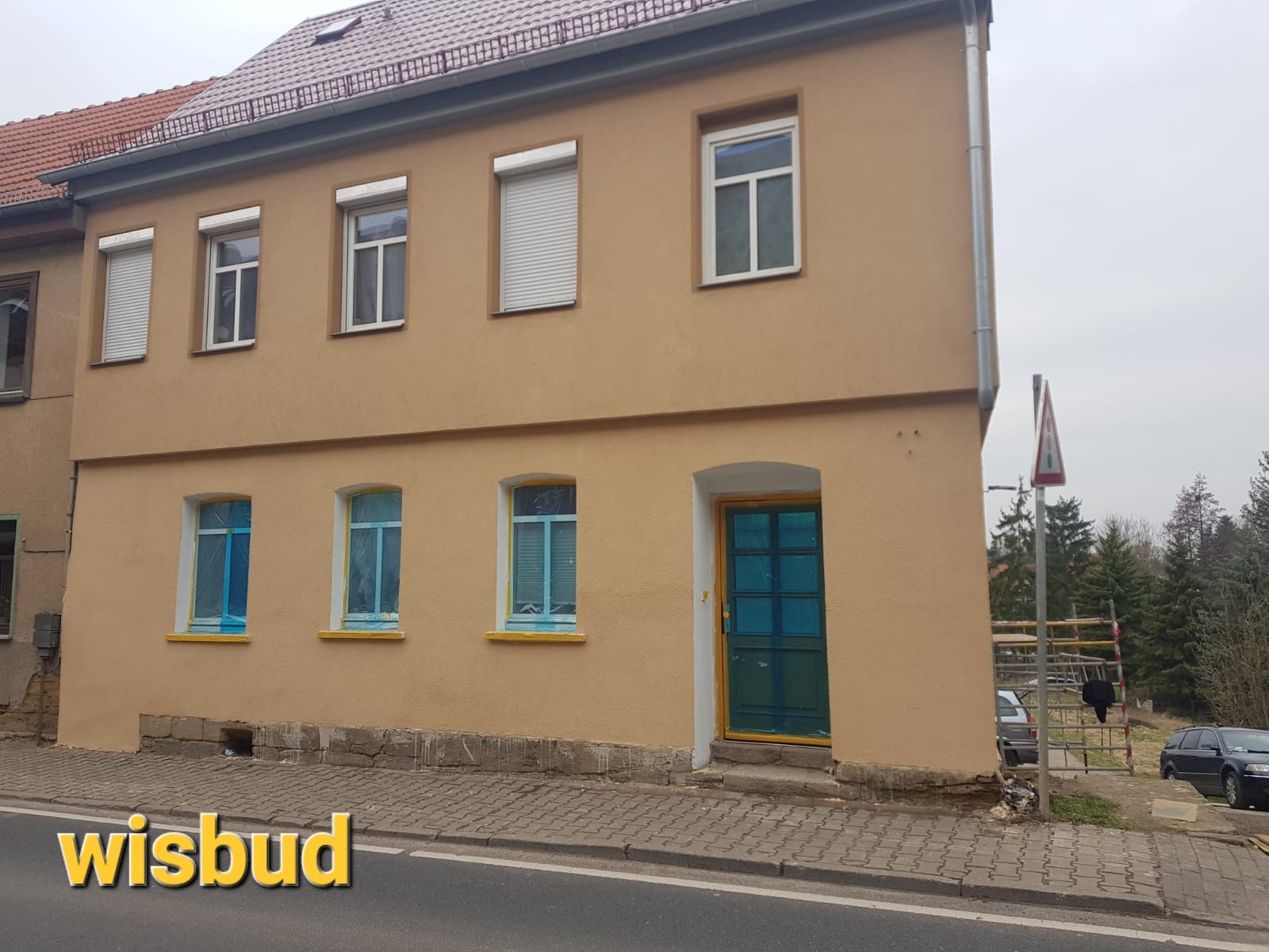 Dwupiętrowy budynek w trakcie prac elewacyjnych: okna i drzwi zabezpieczone folią, świeżo pomalowana fasada w odcieniach beżu, widoczne rusztowania i znak drogowy w tle.