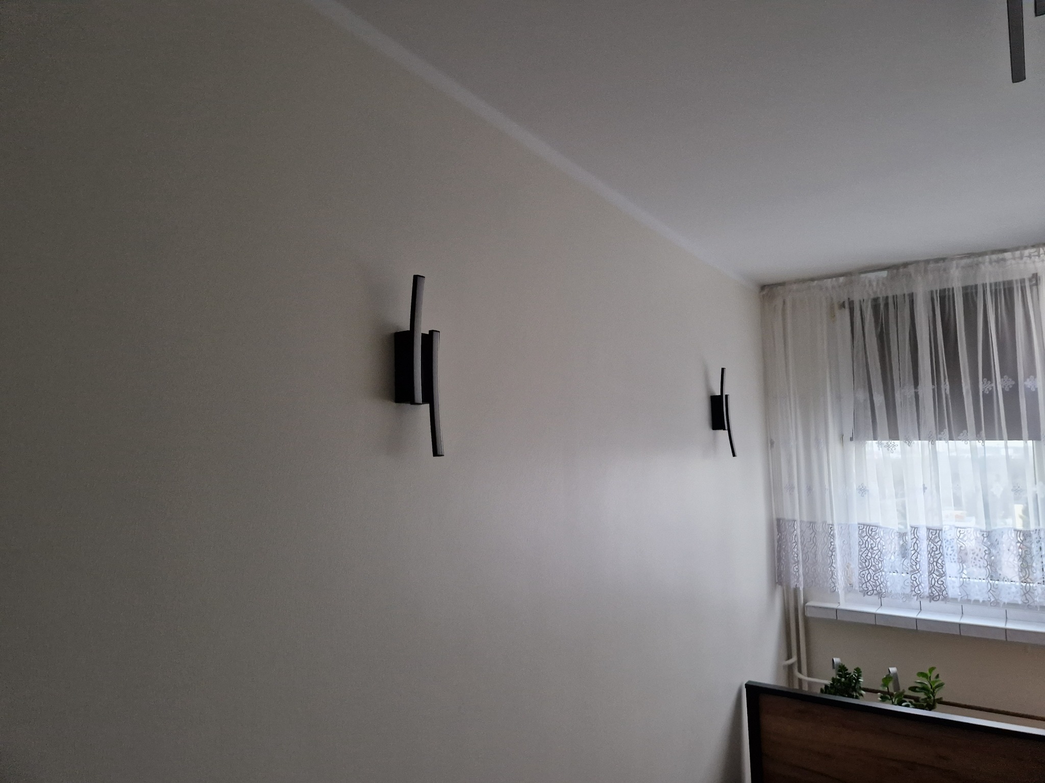Dwa minimalistyczne, czarne kinkiety ścienne zamontowane na jasnoszarej ścianie w pokoju z oknem zasłoniętym firanką. Widoczny fragment ramy łóżka i doniczki z roślinami.