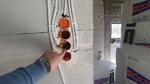 Wykonywanie instalacji elektrycznej w ścianie z betonu komórkowego. Widoczne puszki instalacyjne w trakcie montażu, przewody elektryczne i dłoń wskazująca na jedną z puszek. W tle surowy stan budynku.