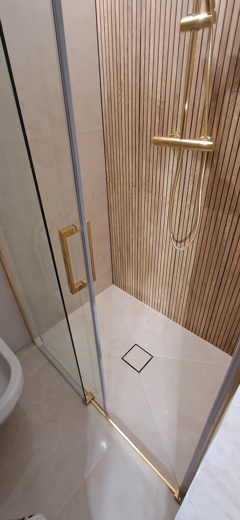 Kabina prysznicowa walk-in z złotymi profilami i deszczownicą na tle ściany z drewnianych paneli, minimalistyczny odpływ liniowy w posadzce.