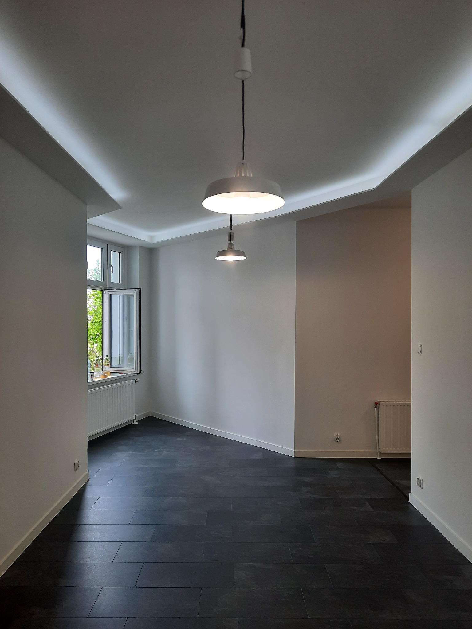 Pusty pokój z ciemną podłogą z płytek, białymi ścianami, oknem z widokiem na zieleń i dwoma wiszącymi lampami z białymi kloszami, oświetlenie LED w suficie.