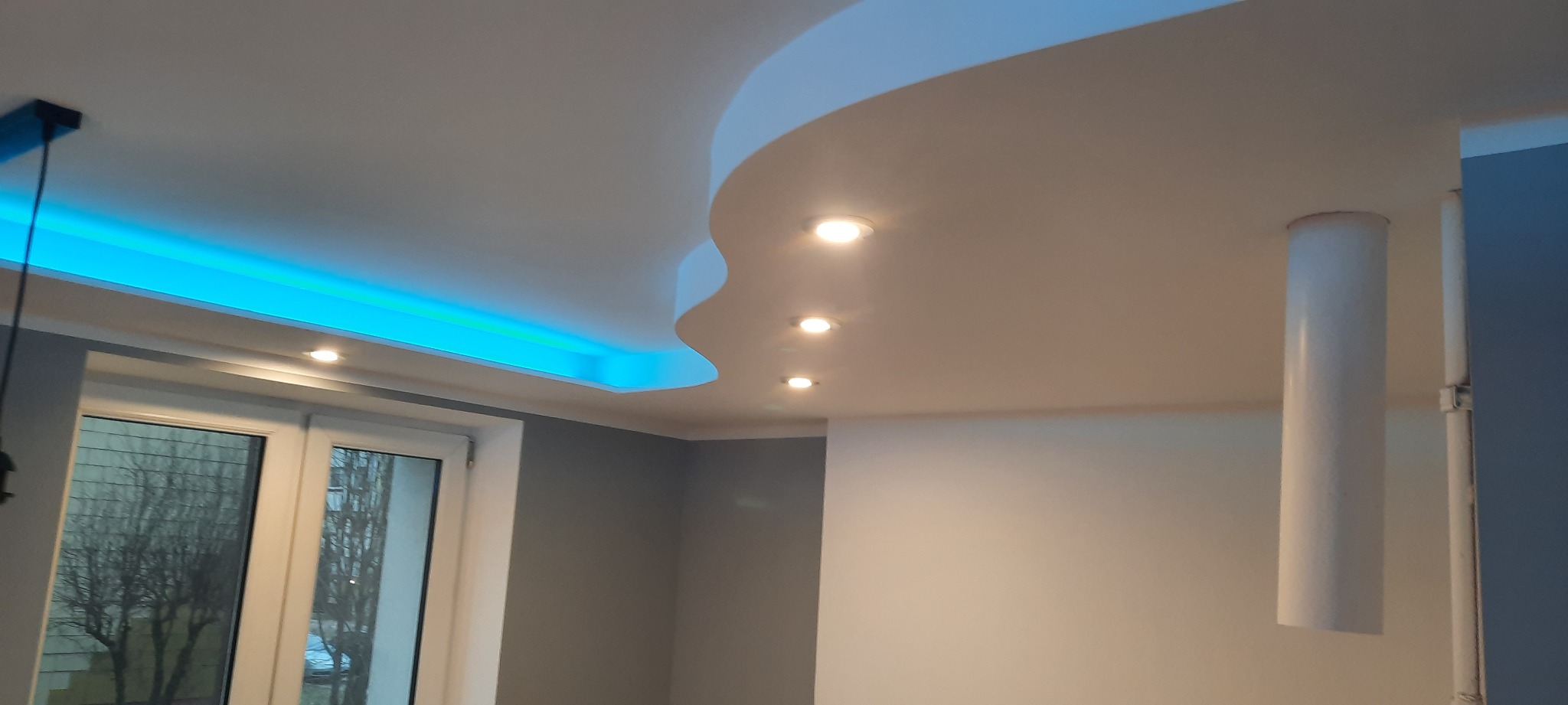 Nowoczesny sufit podwieszany z oświetleniem LED w kolorze niebieskim i wbudowanymi punktami świetlnymi, widoczny fragment okna z widokiem na drzewa.