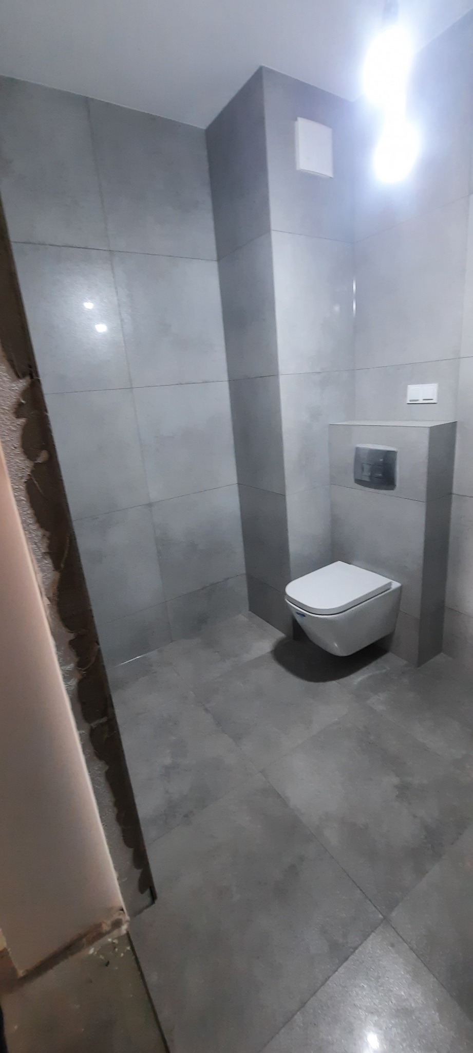 Szara łazienka z wiszącą toaletą, widoczne płytki na ścianach i podłodze, zarys niedokończonego wykończenia po lewej stronie.