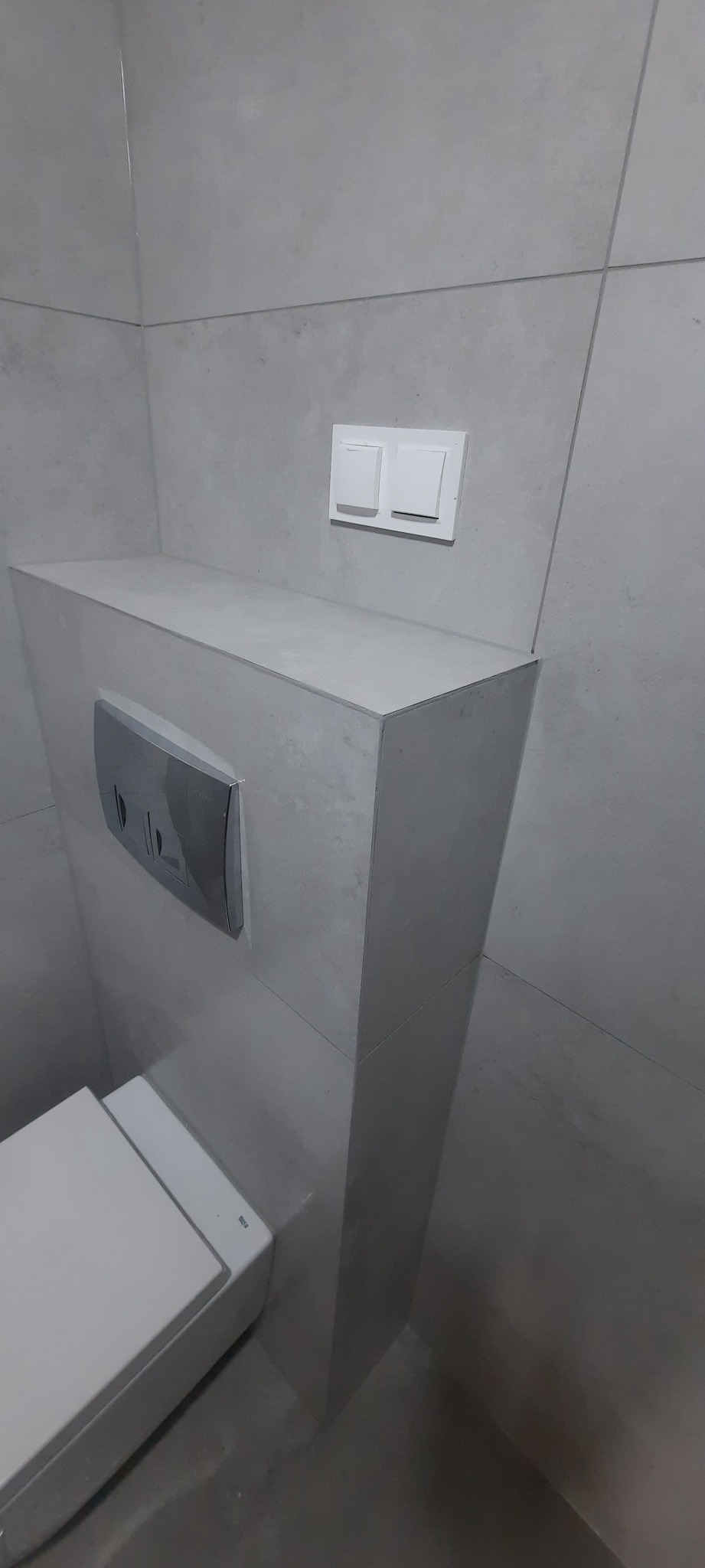 Szara łazienka z zabudowaną spłuczką i przyciskiem, widoczny fragment toalety, nowoczesne wykończenie.