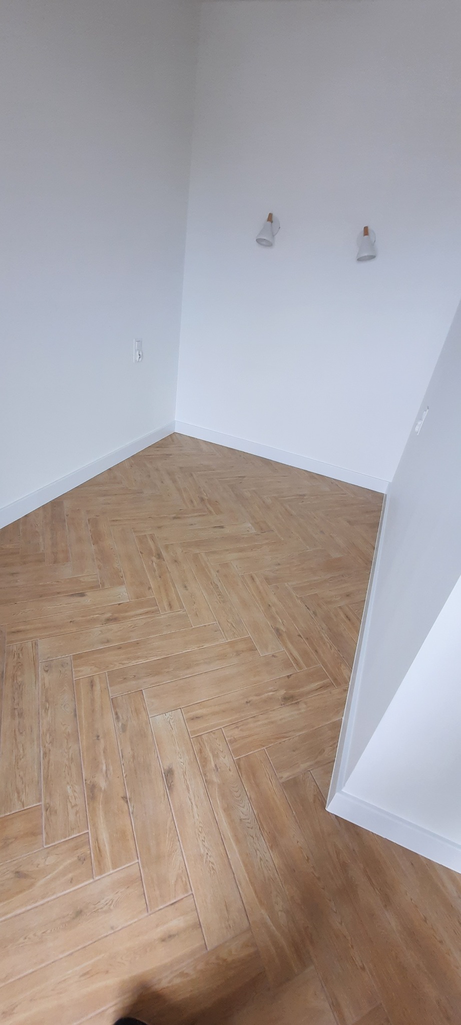 Ukończone podłogi z płytek imitujących drewno ułożonych we wzór jodełki w nowym, pustym pokoju z białymi ścianami i listwami przypodłogowymi, widoczne dwa kinkietowe oświetlenia na ścianie.