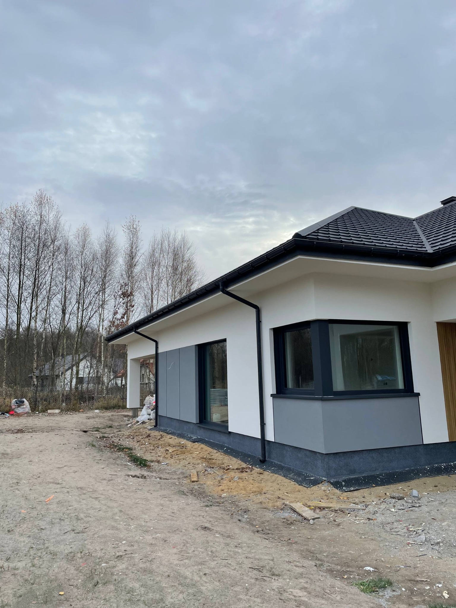 Nowoczesny dom jednorodzinny z białą elewacją i szarymi akcentami, czarną stolarką okienną i dachem. Widoczne elementy wykończeniowe i teren wokół domu w trakcie zagospodarowania.