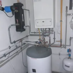 Kompletna instalacja grzewcza z piecem gazowym, zasobnikiem ciepłej wody, pompą obiegową i naczyniem przeponowym, rury miedziane i plastikowe, widoczne zawory kulowe i manometry.