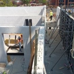 Quick Build - Budowa domu z białych bloczków: widok z góry na wznoszone ściany i szalunki fundamentowe, pracownicy w kaskach na placu budowy.