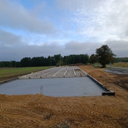 Quick Build - Wylany fundament pod budowę domu jednorodzinnego, widoczne zbrojenia i szalunki, w tle betonowe bloczki i betoniarka, krajobraz wiejski.