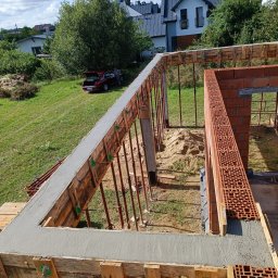 Quick Build - Wylany betonowy wieniec w szalunku, widoczne podpory i fragment muru z czerwonej cegły, w tle domy i samochód osobowy