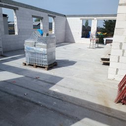 Quick Build - Wnętrze surowego domu murowanego w trakcie budowy, widoczne ściany z białych bloczków, otwory okienne, paleta z materiałami budowlanymi i czerwone rusztowania w rogu.