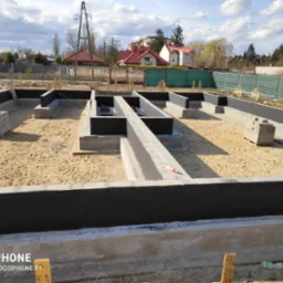 Zalane fundamenty domu z hydroizolacją, widoczne zbrojenia i bloczki betonowe na placu budowy w słoneczny dzień.