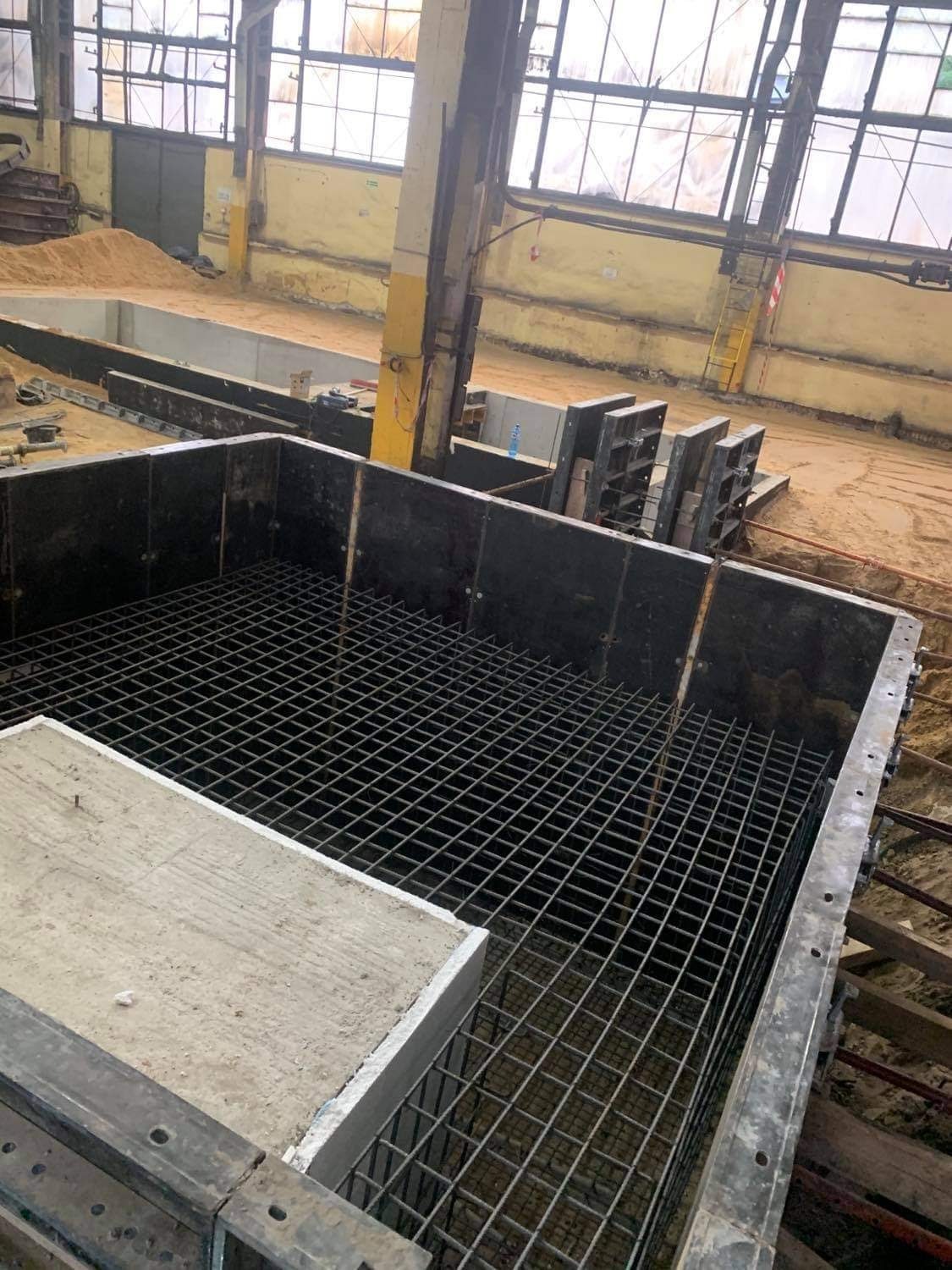 Zbrojenie w szalunku fundamentu, widoczne metalowe pręty tworzące siatkę, fragment betonowej płyty i ściany szalunku, w tle hala budowlana z oknami.
