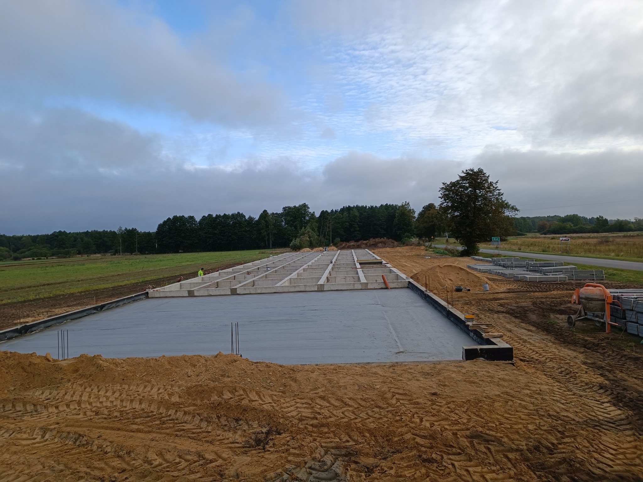 Wylany fundament pod budowę domu jednorodzinnego, widoczne zbrojenia i szalunki, w tle betonowe bloczki i betoniarka, krajobraz wiejski.